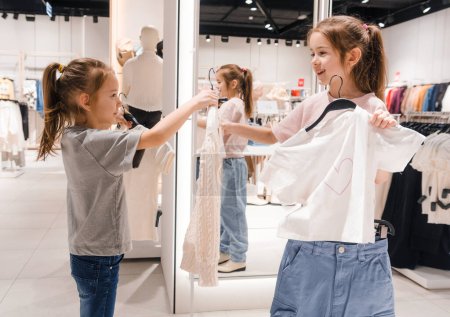 Deux jeunes filles comparent volontiers les choix de vêtements dans un magasin de vêtements lumineux, s'engagent les unes avec les autres et profitent de l'expérience d'achat.