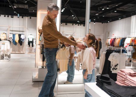 Un père et sa fille partagent un moment de collage tout en magasinant pour des vêtements dans un magasin lumineux, entouré de rayonnages de vêtements.
