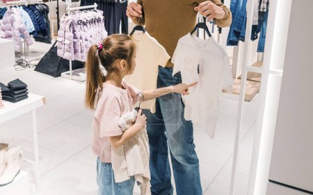Ein junges Mädchen zeigt auf ein Hemd in einem Bekleidungsgeschäft, während ihre Eltern ihr zwei Möglichkeiten zur Auswahl vorhalten.