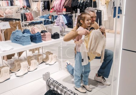 A father helps his excited daughter choose clothes in a brightly lit mall, surrounded by folded clothing and footwear.