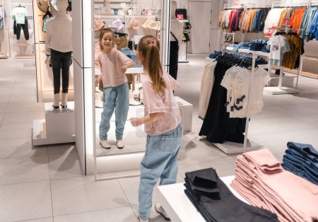 Une jeune fille avec une queue de cheval tente joyeusement ses vêtements tout en se regardant dans un miroir pleine longueur à l'intérieur d'un magasin de vêtements soigneusement arrangé et bien éclairé.