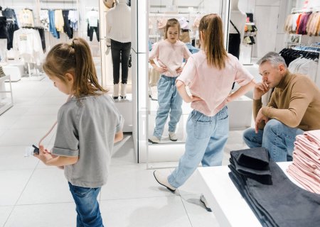 Téléchargez les photos : Un père s'assoit soigneusement tandis que ses deux filles essaient de nouvelles tenues dans un magasin de vêtements moderne, entouré de rayonnages de vêtements élégants. - en image libre de droit