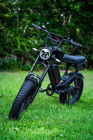 Foto de Una elegante bicicleta eléctrica negra, que recuerda a un ciclomotor, se levanta con gracia en un paisaje herboso sereno, rodeado de árboles - Imagen libre de derechos