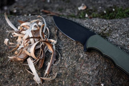 Téléchargez les photos : Un couteau de poche noir et vert assis à côté de lambeaux de bois pour allumer un feu dans la nature sauvage - en image libre de droit