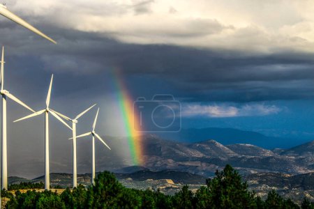 Photo for Picturesque scenery of modern white windmills located in scenic mountainous countryside against fluffy gray clouds floating in blue sky with bright rainbow after rain - Royalty Free Image