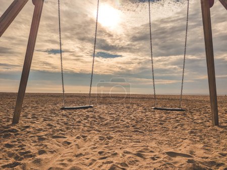 Balançoires suspendues à des poteaux en bois érigés sur une plage de sable avec des chaînes en plein soleil contre des nuages recouvrant le soleil brillant dans le ciel bleu en journée