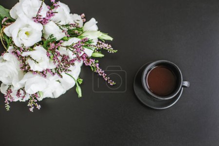 Black ceramic cup with coffee on table and bouquet of flowers. Copy space.
