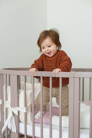 Téléchargez les photos : Cute laughing baby standing in round bed. Little girl learns to stand in her crib. - en image libre de droit