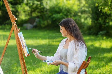 Téléchargez les photos : Young female artist paints picture in summer park in sunlight. Art and hobby concept. - en image libre de droit