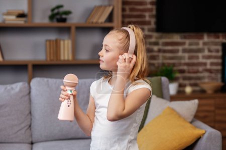 Photo for Happy little girl listening to music at home. Child wearing headphones using microphone, singing song on sofa in room. - Royalty Free Image