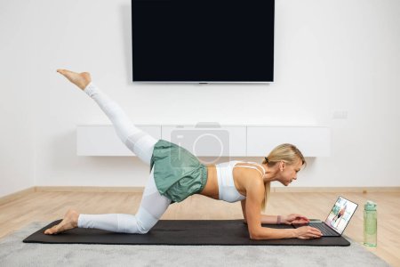 Training Concept. Fit woman doing yoga plank and watching online tutorials on laptop, training in room.