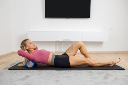 Fit athletic woman in sportswear ride on foam myofascial massage roller, massaging their muscles on floor of yoga mat in home. MFR