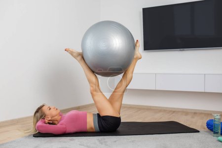 Millennial-Frau trainiert mit Fitnessball auf Gymnastikmatte im Wohnzimmer. Fitness, Sport und gesunder Lebensstil.