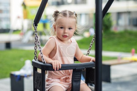 Kid toddler girl swinging on playground swing. Kid playing on school or kindergarten yard. Summer time.
