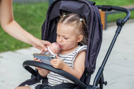 Téléchargez les photos : Petit enfant en poussette sirote de bouteille tout en regardant curieusement smartphon - en image libre de droit