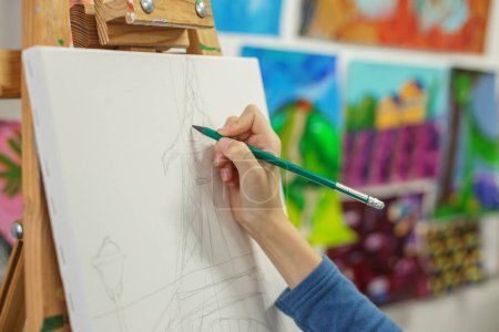 Nahaufnahme der Hand eines Künstlers mit Bleistift, Zeichnung vorläufiger Linien für Skizzen auf weißer Leinwand mit farbenfrohen Gemälden im Hintergrund.