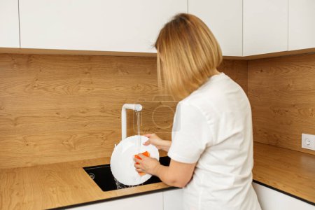 Hände waschen einen weißen Teller unter fließendem Wasser in einer glatten schwarzen Küchenspüle und präsentieren alltägliche Haushaltsarbeiten.