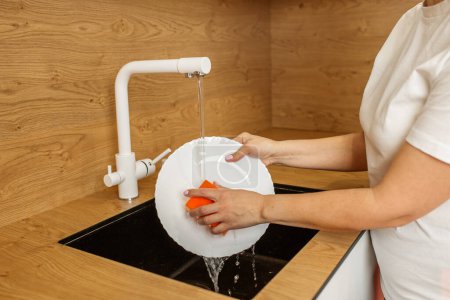 Hands washing a white plate under running water in a sleek black kitchen sink, showcasing everyday household tasks.
