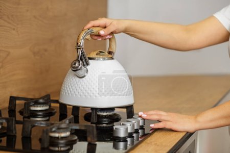 Person's hand is placing stylish white kettle on stove top, preparing for hot beverage in cozy kitchen atmosphere.