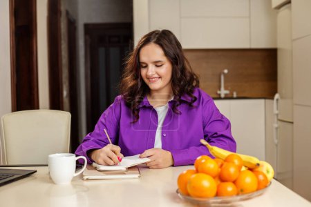 Cheerful woman in purple shirt is jotting down notes in notebook at her kitchen table, with cup of coffee and bowl of fruit adding to pleasant atmosphere.