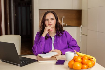 Nachdenkliche Studentin sitzt an ihrem Küchentisch mit Laptop, Notebook und Smartphone und plant ihren Tag bei einer Tasse Kaffee und einer Schüssel mit frischem Obst
