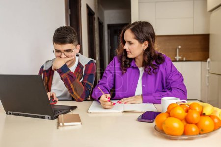 Teenage girl and boy concentrate on their homework, using a laptop and taking notes at a kitchen table, collaborating in home environment.