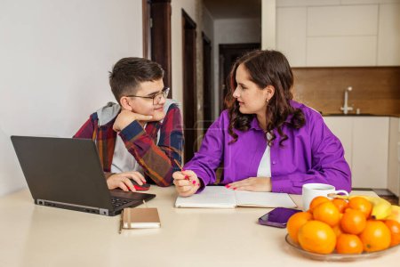 Teenage girl in purple shirt writes in notebook, discussing academic work with teen boy in glasses, with laptop open in front of them.