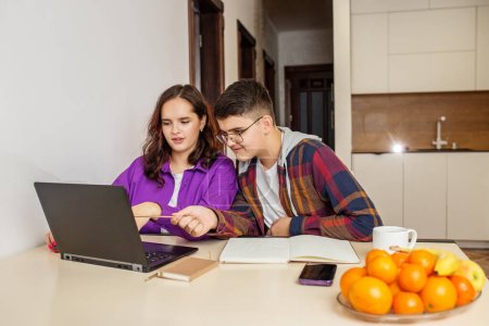 Two teenage students, girl and boy, are engaging in a group study session with a laptop, books, and stationery on kitchen table.
