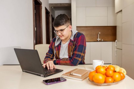 Focused teenage boy with glasses is intently studying on his laptop, surrounded by study materials and fresh fruit on table.