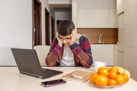 Teenage boy wearing glasses looks stressed while studying on his laptop, with fruit and coffee mug on table. Back to school.