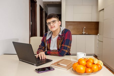 Teen boy wearing glasses is concentrating on his work using a laptop, with smartphone, notebook, and healthy fruits on table. Back to school.