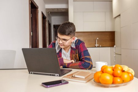 Foto de Enfocado adolescente con gafas está estudiando intensamente en su ordenador portátil, rodeado de materiales de estudio y fruta fresca en la mesa. General Z. - Imagen libre de derechos
