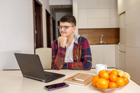 Foto de Enfocado adolescente con gafas está estudiando intensamente en su ordenador portátil, rodeado de materiales de estudio y fruta fresca en la mesa. General Z. - Imagen libre de derechos
