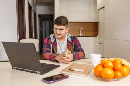 Foto de Enfocado adolescente con gafas está estudiando intensamente en su ordenador portátil, rodeado de materiales de estudio y fruta fresca en la mesa - Imagen libre de derechos