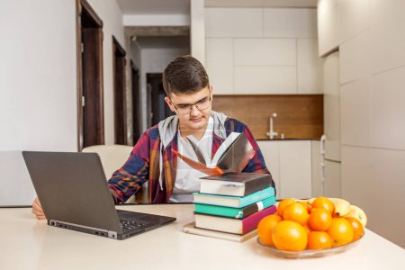 Young student in glasses is deeply focused on reading a book while working with laptop, surrounded by stack of books and fresh fruit. Back to school. Gen Z.