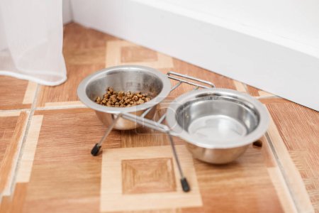 Two stainless steel pet bowls on wooden floor, one with food and one empty, suggesting feeding area for pet.