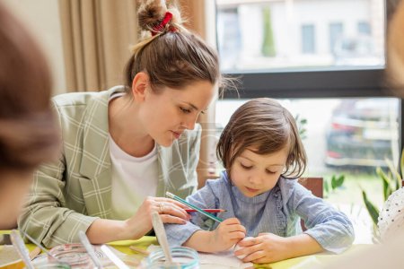 Mother attentively guides little daughter in art of Easter egg painting at home.