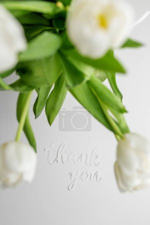 White tulips and 'thank you' cutout create an elegant expression of gratitude against soft grey background.