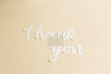 High-angle view of thank you in white cutout letters against a smooth beige background, portraying message of thanks.