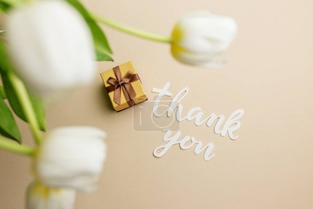 Heartfelt thank you message in cursive script, accompanied by small gift box and soft white tulips on beige background.
