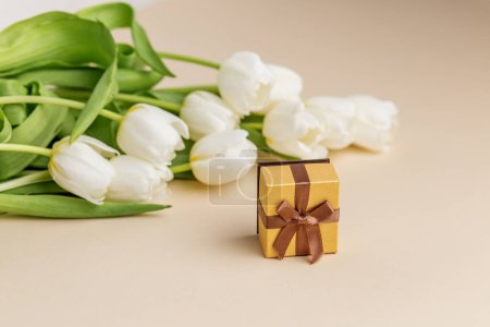 Geschmackvolle Komposition mit weißen Tulpen neben einem charmanten Geschenkkarton mit braunem Band vor neutralem beigem Hintergrund.
