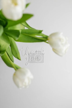 White tulips and 'thank you' cutout create an elegant expression of gratitude against soft grey background.