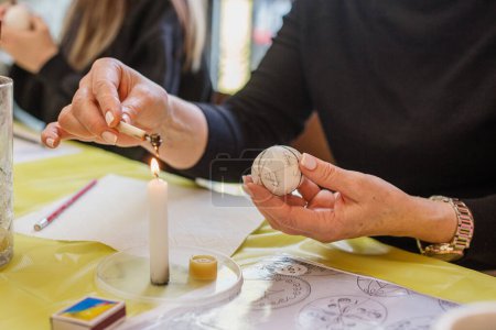 Hand hält ein Wachswerkzeug über eine Kerzenflamme und zeichnet vorsichtig Muster auf ein Osterei.