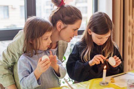 Tierno momento familiar como madre enseña a sus dos hijas el arte de la decoración de huevos de Pascua en su casa.