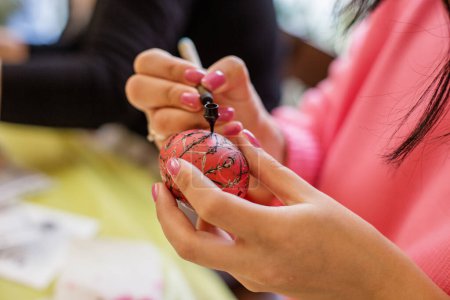 Close-up of hands applying black wax patterns on a vibrant red Easter egg, a beautiful traditional art form.