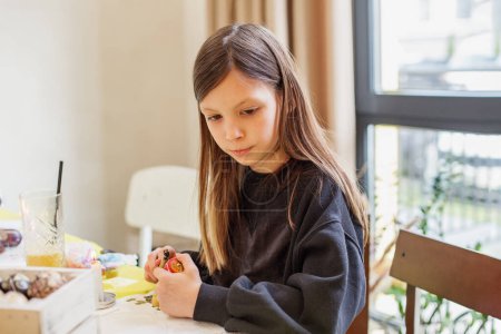 Child girl with brown hair is deeply concentrated on painting a colorful Easter egg, embodying the spirit of holiday.