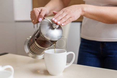 Freshly brewed tea is being poured from a stainless steel French press into white mug