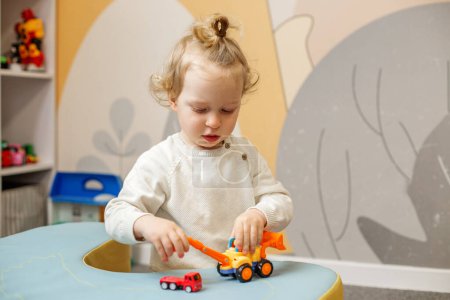 Intuitiv fokussierter Kleinkind greift mit Spielzeugfahrzeugen auf Spieltisch in gut organisiertem Kinderzimmer ein.