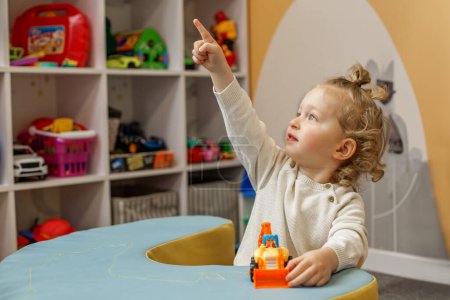Adorable toddler boy with toy pointing up while sitting at play table in colorful playroom.