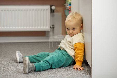 A relaxed toddler with a contemplative expression sitting on carpet in a cozy corner of room.